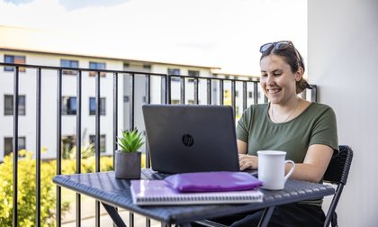 A student sitting on a balcony with their laptop. They are smiling at the screen.