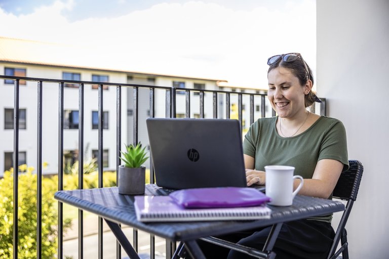 A student sitting on a balcony with their laptop. They are smiling at the screen.
