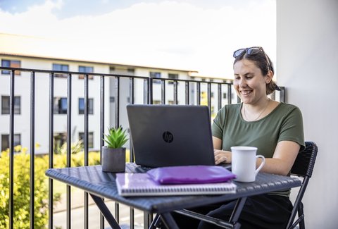 A student sitting on a balcony with their laptop. They are smiling at the screen.