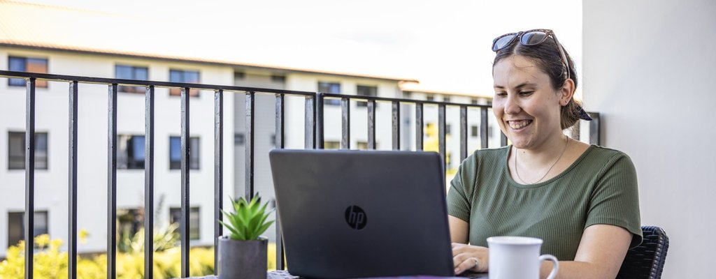 A student sitting on a balcony with their laptop. They are smiling at the screen.