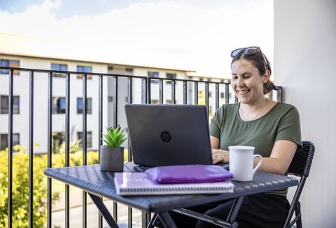 A student sitting on a balcony with their laptop. They are smiling at the screen.