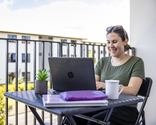 A student sitting on a balcony with their laptop. They are smiling at the screen.
