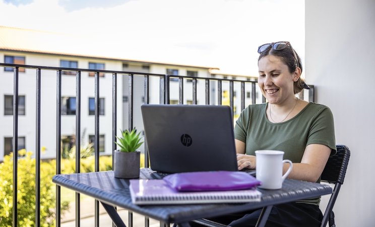 A student sitting on a balcony with their laptop. They are smiling at the screen.