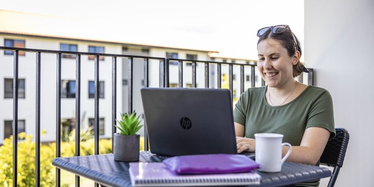 A student sitting on a balcony with their laptop. They are smiling at the screen.